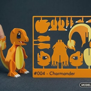 Könnte beinhalten: Ein orange-gelber Kunststoff-Modellbausatz der Figur Glumanda, bestehend aus einer fertig montierten Figur und einem flachen Rahmen mit verschiedenen Einzelteilen. Der Rahmen ist mit #004 - Charmander beschriftet und enthält ein Model Kit Logo.