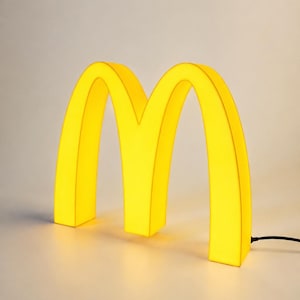 Könnte beinhalten: Eine leuchtend gelbe, beleuchtete Leuchte in Form des Buchstabens M. Die Struktur weist ein glattes, geschwungenes Design mit einer durchscheinenden Oberfläche auf, die von innen leuchtet. Ein dünnes schwarzes Stromkabel führt aus der Basis.