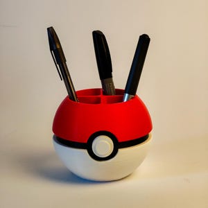 Puede incluir: Un organizador de escritorio esférico diseñado para parecerse a una bola roja y blanca con una banda horizontal negra y un botón central blanco. La parte superior tiene cuatro compartimentos con tres bolígrafos negros.