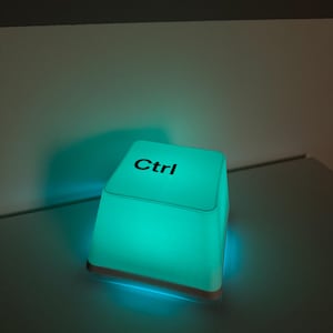 Può includere: Una lampada quadrata a forma di tasto di computer, realizzata in materiale bianco traslucido che emette una luminosa luce color ottanio. La superficie superiore della lampada presenta la scritta nera Ctrl in carattere sans-serif.