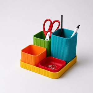 Può includere: Un organizzatore da scrivania a più scomparti con un vassoio base giallo, un contenitore quadrato arancione, uno verde e uno alto color ottanio. All'interno sono riposte forbici rosse, una penna nera, una penna bianca e alcune graffette argentate.