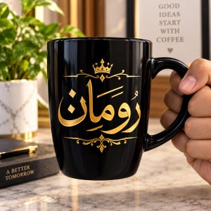 Könnte beinhalten: Eine glänzend schwarze Keramiktasse mit goldener arabischer Kalligrafie in der Mitte, gekrönt von einem kleinen goldenen Kronensymbol und eingerahmt von dekorativen goldenen Verzierungen. Das Design ist auf der Vorderseite der Tasse gedruckt.