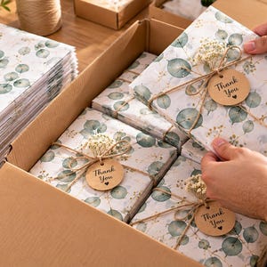 May include: Rectangular packages wrapped in white paper with a green eucalyptus leaf pattern, tied with brown twine. Each package features a small, round, tan cardboard tag with the text Thank You and a heart symbol, accented by dried flowers.