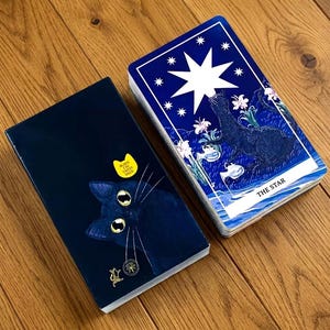 May include: A deck of tarot cards featuring a dark blue box with a black cat illustration and a yellow sticker reading Black Cat Tarot Deck. Beside it, a card displays a large white star, a black cat, and flowers with the text The Star.
