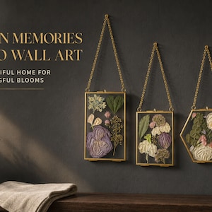 Può includere: Tre cornici in vetro sospese con bordi in metallo dorato, ciascuna contenente fiori secchi pressati. Le cornici sono appese tramite catenelle dorate. Il testo sulla parete recita: TURN MEMORIES INTO WALL ART. A BEAUTIFUL HOME FOR MEANINGFUL BLOOMS.