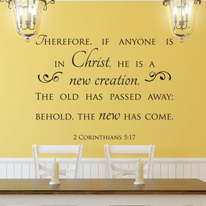 Puede incluir: Una pared amarilla con un zócalo blanco. Una calcomanía de pared de vinilo negro con un versículo bíblico de 2 Corintios 5:17 dice "Por lo tanto, si alguno está en Cristo, nueva criatura es; las cosas viejas pasaron; he aquí, todas son hechas nuevas." La calcomanía está sobre una mesa de madera con sillas blancas.