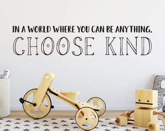 Peut inclure: Un mur blanc avec une citation inspirante en noir et blanc qui dit "Dans un monde où vous pouvez être n'importe quoi, choisissez la gentillesse". Le mur est décoré d'un tricycle en bois, d'un robot en bois et d'un panier en osier avec des animaux en peluche.