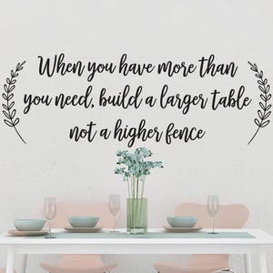 May include: A white wall with black calligraphy that reads "When you have more than you need, build a larger table not a higher fence". The wall is decorated with a vase of light blue flowers and a white table with pink chairs.