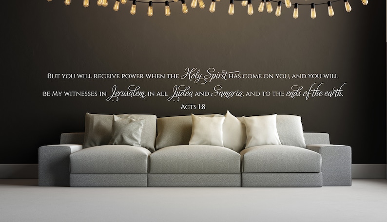 May include: A living room with a gray couch and white throw pillows. The wall behind the couch has a white vinyl decal with a bible verse from Acts 1:8, "But you will receive power when the Holy Spirit has come on you, and you will be my witnesses in Jerusalem, in all Judea and Samaria, and to the ends of the earth."