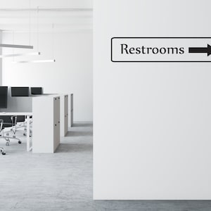 May include: A white wall with a black sign that says "Restrooms" with a black arrow pointing to the right.