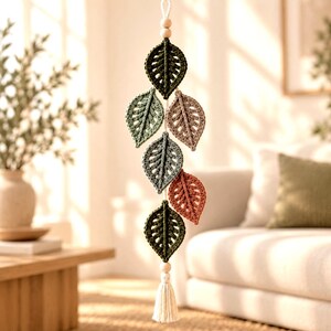 Puede incluir: Un colgante de pared vertical con cinco hojas de ganchillo en tonos verde oscuro, verde salvia, rosa empolvado, azul pizarra y terracota. Las hojas están ensartadas en un cordón con dos cuentas de madera clara arriba y una borla color crema.