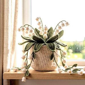 Puede incluir: Una planta de lirio de los valles tejida a ganchillo con flores blancas en forma de campana y hojas verdes texturizadas. La planta está en una maceta cilíndrica de ganchillo beige con un patrón de puntos visible, sobre una superficie de madera.