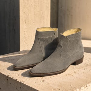 May include: A pair of grey suede ankle boots with a pointed toe, a low stacked heel, and a dark brown sole. The boots feature a side panel with vertical stitching details and a light-coloured interior lining, showcasing a clean and structured design.