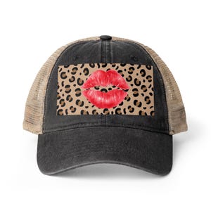May include: A black trucker hat featuring a rectangular patch on the front panel. The patch displays a tan and brown leopard print pattern with a pair of bright red lips centered in the middle. The hat has a curved brim and tan mesh back.