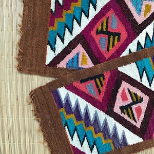 Op de afbeelding: Twee rechthoekige geweven textielstroken met een bruine rand en een centraal patroon van ruiten en zigzags in levendige tinten magenta, turkoois, geel, lichtroze, paars en wit, met zichtbare gerafelde randen aan de zijkanten.