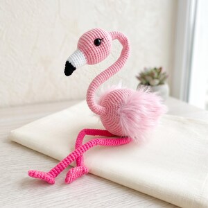 May include: A crocheted flamingo toy featuring a light pink yarn body with a long curved neck, a white and black beak, and dark plastic eyes. The flamingo has long, thin, bright pink crocheted legs and soft, fluffy pink faux fur accents.