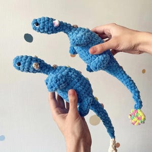 May include: Two blue crochet dinosaur plush toys with long necks and tails. Each dinosaur features a textured yarn body, small dark eyes, and a multi-colored pom-pom or tuft of yarn at the end of the tail. The creatures have small limbs and a rounded head.