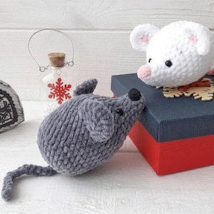 May include: Two crocheted plush mice, one white with a pink nose and one gray with a black nose, are positioned near a red and blue gift box. A glass jar with a red snowflake ornament and a heart-shaped wooden decoration are also visible.