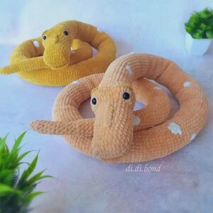 Puede incluir: Dos serpientes de peluche enrolladas hechas de hilo texturizado. Una serpiente es de color amarillo brillante y la otra es de un color melocotón más claro, ambas con manchas blancas y ojos de cuentas negras. El texto di.di.bond es visible abajo.
