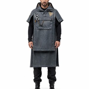 Könnte beinhalten: Ein dunkelgrauer Kapuzenponcho mit einer großen Vordertasche, einem silberfarbenen Karabinerhaken und einem dreieckigen Aufnäher auf der Brust. Das Kleidungsstück hat kurze Ärmel, Metallösen am Saum und eine lange, strukturierte Silhouette.