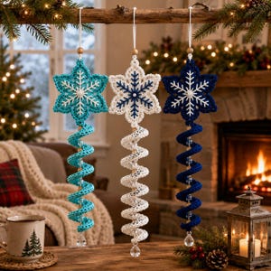 May include: Three hanging crochet ornaments featuring six-pointed snowflake tops in turquoise, white, and navy blue. Each snowflake is attached to a spiraling, corkscrew-shaped tail in a matching colour, accented with clear beads and a crystal drop.