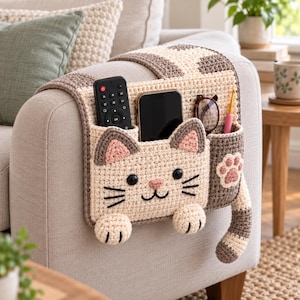 May include: A beige and brown crochet armchair caddy featuring a cat face design with black eyes, a pink nose, and whiskers. The organizer has three pockets holding a black remote, a smartphone, eyeglasses, and a pink crochet hook.