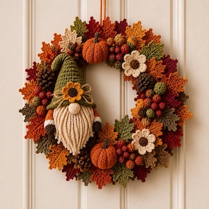 May include: A circular wreath crafted from crocheted autumn leaves in shades of orange, red, brown, and green. The wreath features two crocheted pumpkins, two cream-colored flowers, clusters of red berries, pinecones, and a central gnome figure.