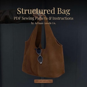 Puede incluir: Un bolso de ante marrón con unas gafas de sol negras colgadas de la correa. El texto dice: Structured Bag, PDF Sewing Pattern & Instructions by Artisan Goods Co. y Premium Suede. El bolso tiene una abertura ancha y un diseño sencillo.
