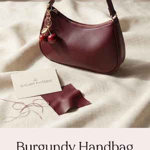Op de afbeelding: Een bordeauxrode handtas met een getextureerd oppervlak, een enkele schouderband en een goudkleurige ritssluiting. Een goudkleurige kersenbedel hangt aan de band. Een klein kaartje bevat de tekst ELEGANT PATTERNS, naast een bordeauxrode stofstaal.