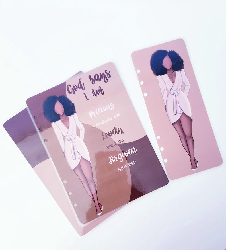May include: Planner inserts with illustrations of a woman in a white dress. The inserts have the text "God says I Am" and the words "Precious," "Lovely," and "Forgiven." The background is a mix of brown and pink.