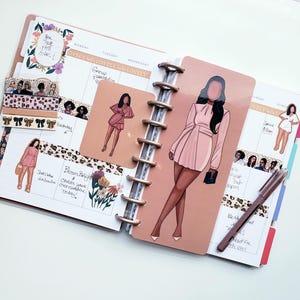 May include: A planner with a spiral binding, open to a spread with illustrations of women in pink dresses. Decorative stickers and washi tape with floral and leopard print patterns are also visible. A pen rests on the right page.