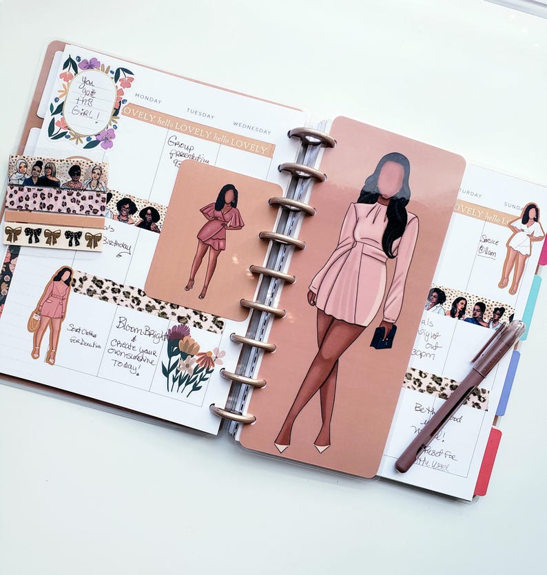 May include: A planner with a spiral binding, open to a spread with illustrations of women in pink dresses. Decorative stickers and washi tape with floral and leopard print patterns are also visible. A pen rests on the right page.