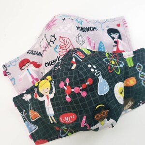 May include: Two fabric face masks with a science theme. One mask is pink with white and black illustrations of scientists, test tubes, and the words "Chemistry" and "Astronomy". The other mask is dark green with white and black illustrations of scientists, test tubes, molecules, and the words "E=MC2".