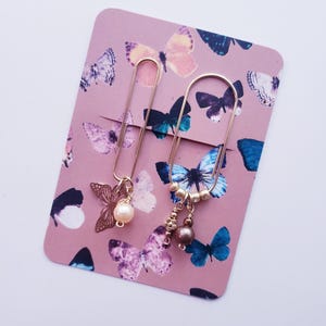 May include: Two gold-colored paper clips with decorative charms on a pink card. One clip has a butterfly charm and a pearl, the other has beads. The card is decorated with butterfly illustrations in shades of purple and blue.