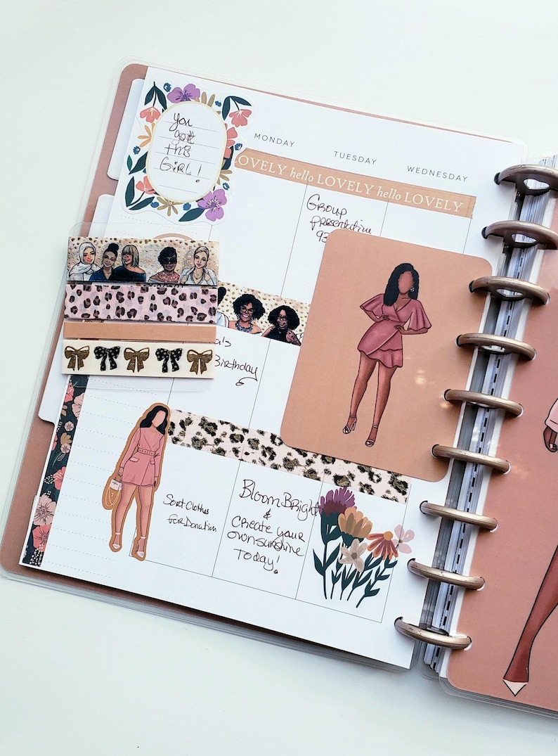 May include: A planner with a pink cover and gold rings, open to a spread with stickers, washi tape, and handwritten notes. The stickers feature illustrations of women in pink dresses. The washi tape has a leopard print and bow design.