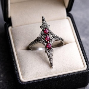 Op de afbeelding: Een zilverkleurige metalen ring met een langwerpig, puntig verticaal ontwerp met gedetailleerde texturen. Drie kleine, ovaalvormige, dieprode edelstenen zijn in een verticale rij geplaatst in het midden van de decoratieve voorzijde.
