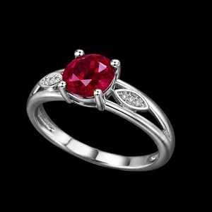 May include: A silver ring featuring a central oval-cut deep red gemstone held by four prongs. The band splits into two arms on each side, with each arm accented by a small leaf-shaped setting containing three tiny clear round-cut stones.