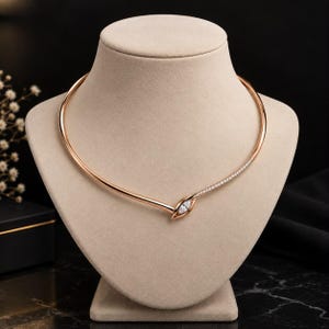 May include: A rose gold-toned collar necklace featuring a smooth, curved band that overlaps at the front. One side is accented with a row of small, clear pavé-set stones, culminating in a single marquise-cut clear stone at the center point.