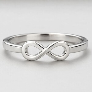 May include: A polished silver-toned ring featuring a central infinity symbol design. The band is smooth and continuous, connecting to the horizontal figure-eight shape at the front. The metal has a reflective, metallic finish throughout the piece.