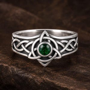 Op de afbeelding: Een zilveren ring met een ingewikkeld gevlochten knooppatroon langs de band en een centrale ronde donkergroene edelsteen in een driehoekig frame. Het metaal heeft een gepolijste afwerking met donkere uitsparingen voor extra detail.