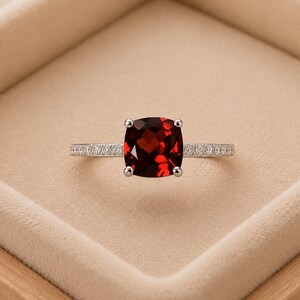 Puede incluir: Un anillo de tono plateado con una piedra preciosa granate de color rojo intenso en talla cojín, sujeta por cuatro garras. La banda delgada está incrustada con una hilera de pequeñas piedras transparentes en talla brillante pavé.