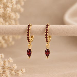 May include: A pair of gold hoop earrings featuring a row of small, round, deep red garnet stones along the front of each hoop. Each earring includes a dangling, marquise-cut, deep red garnet stone suspended from the bottom of the hoop.