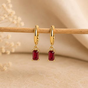 Puede incluir: Un par de pendientes de aro dorados con piedras preciosas rojas rectangulares colgantes. Cada pendiente consta de un aro dorado pulido con una pequeña anilla que conecta a una piedra roja vibrante de corte baguette.