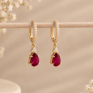 May include: A pair of gold hoop earrings featuring a small cluster of clear stones at the top and a dangling teardrop-shaped red gemstone. The hoops are also accented with tiny clear stones along the front-facing outer edge of the circular band.