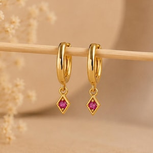Puede incluir: Un par de pendientes de aro dorados con un pequeño dije en forma de diamante colgando de cada aro. Cada dije tiene una piedra preciosa de color rosa vibrante engastada en el centro. Los pendientes cuelgan de un fino palo de madera.