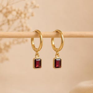 May include: A pair of gold hoop earrings, each featuring a small, rectangular, deep red gemstone charm suspended from the bottom. The earrings are displayed hanging from a thin, light-colored wooden rod, showcasing the polished metal finish.
