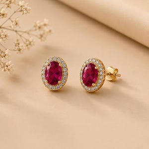 May include: A pair of gold stud earrings featuring oval-cut deep red gemstones at the centre. Each gemstone is surrounded by a halo of small, clear, round-cut white stones set in a gold frame. The earrings include gold posts and secure backings.