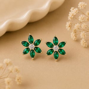 May include: A pair of stud earrings featuring a flower design with six marquise-cut green stones surrounding a single round clear stone in the center. The stones are set in a gold-toned metal frame, creating a symmetrical floral appearance.