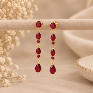 Puede incluir: Un par de pendientes largos de oro con una disposición vertical de tres piedras rojas en forma de pera y dos piedras rojas pequeñas y redondas, unidas por finos eslabones dorados con un acabado de metal dorado pulido.