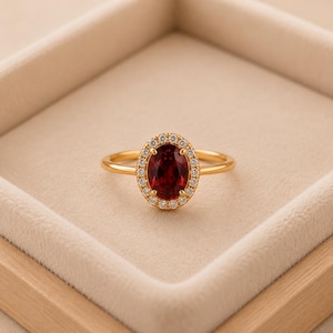 Puede incluir: Un anillo de oro con una piedra preciosa de color rojo intenso de corte ovalado en el centro, rodeada por un halo de pequeñas piedras transparentes de corte redondo. El aro es de oro liso y pulido, con un diseño clásico y una piedra roja.